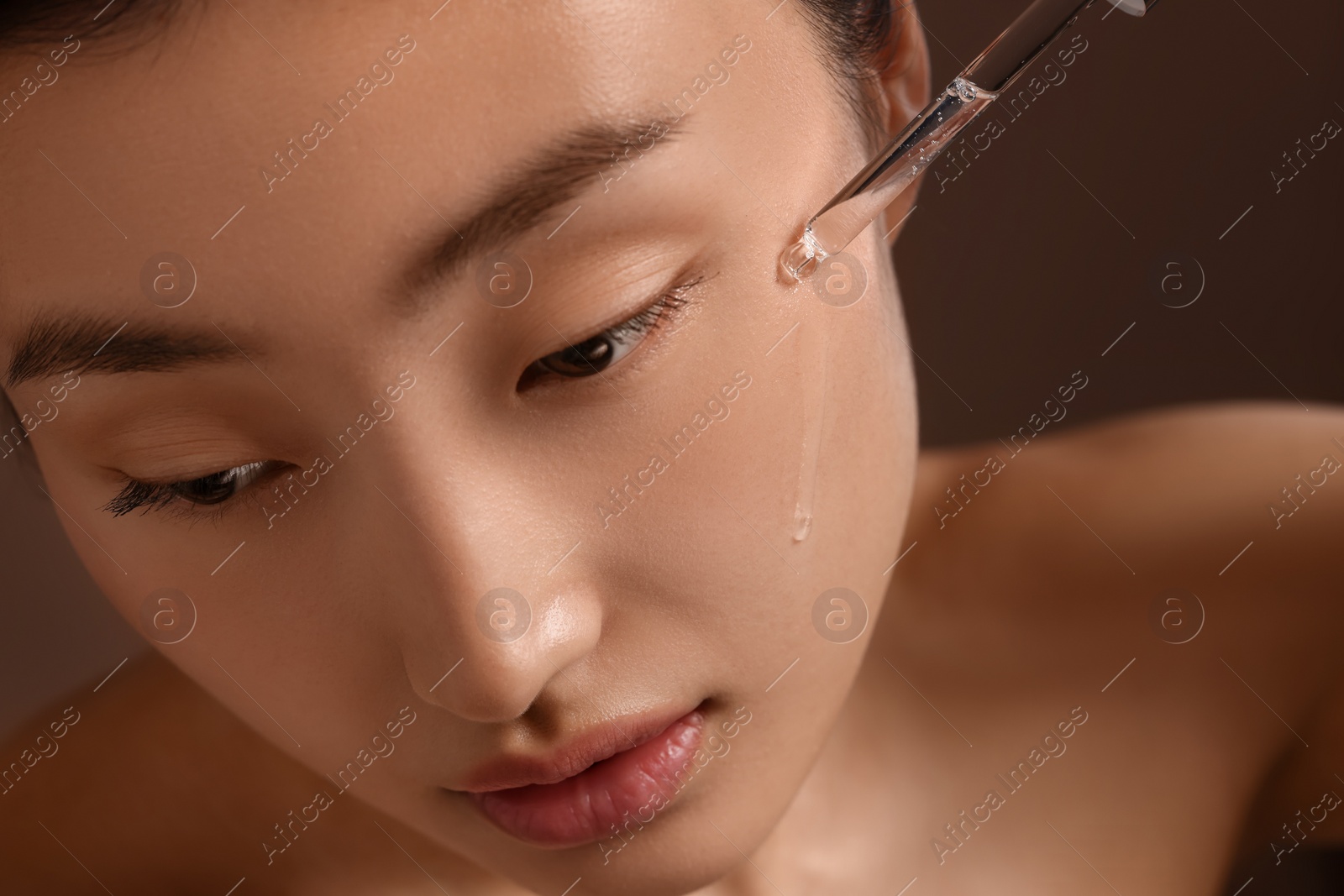 Photo of Beautiful young woman applying cosmetic serum onto her face on brown background, closeup