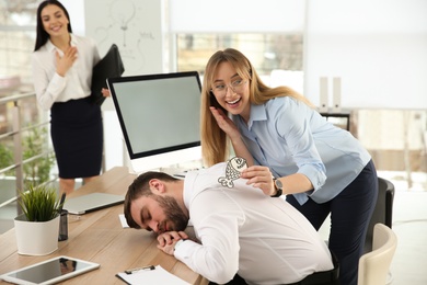 Young woman sticking paper fish to colleague's back while he sleeping in office. Funny joke