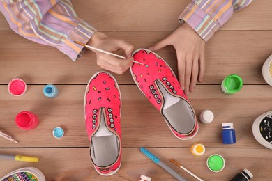 Photo of Woman painting on sneaker at wooden table, top view. Customized shoes