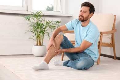 Photo of Man massaging leg on white carpet near armchair at home