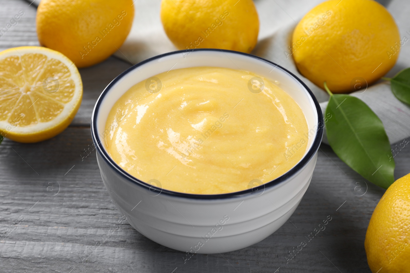 Photo of Delicious lemon curd in bowl, fresh citrus fruits and green leaf on grey wooden table, closeup