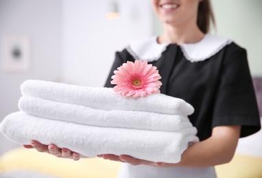 Photo of Young maid holding stack of towels and flower in hotel room