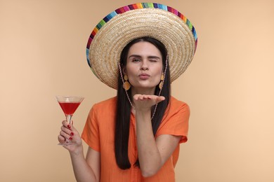 Photo of Young woman in Mexican sombrero hat with cocktail blowing kiss on beige background