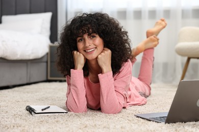 Beautiful young woman in stylish pyjama with laptop and notebook on floor at home