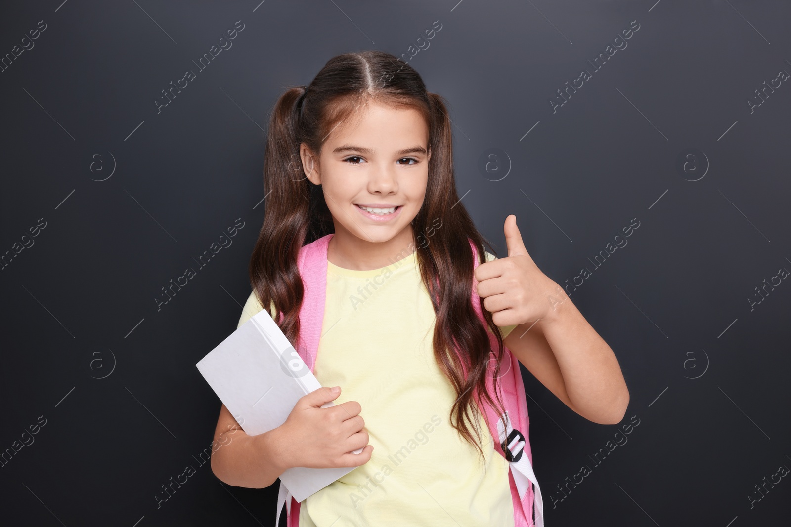 Photo of Cute schoolgirl with book showing thumbs up near chalkboard