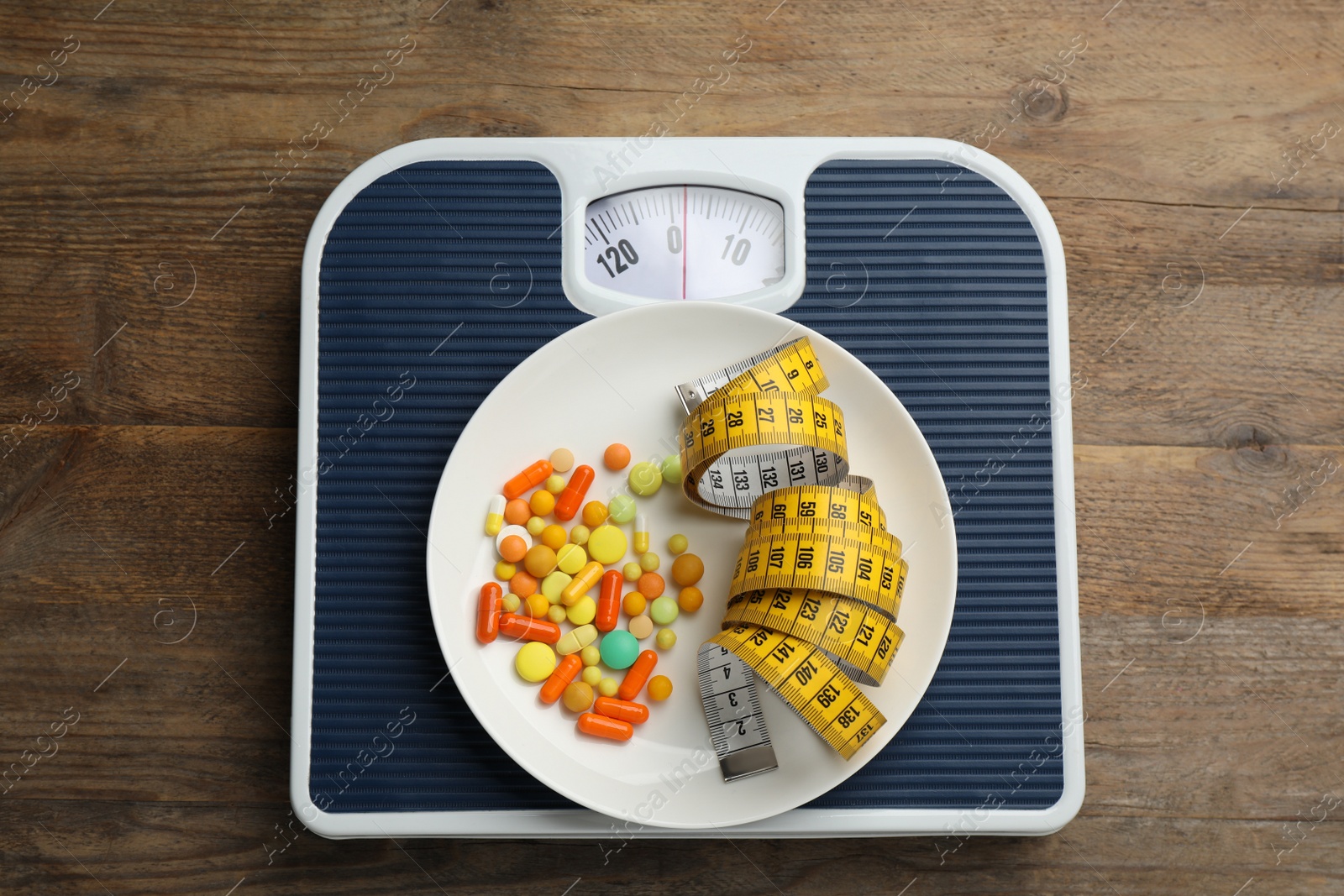 Photo of Scales with different weight loss pills and measuring tape on wooden table, top view