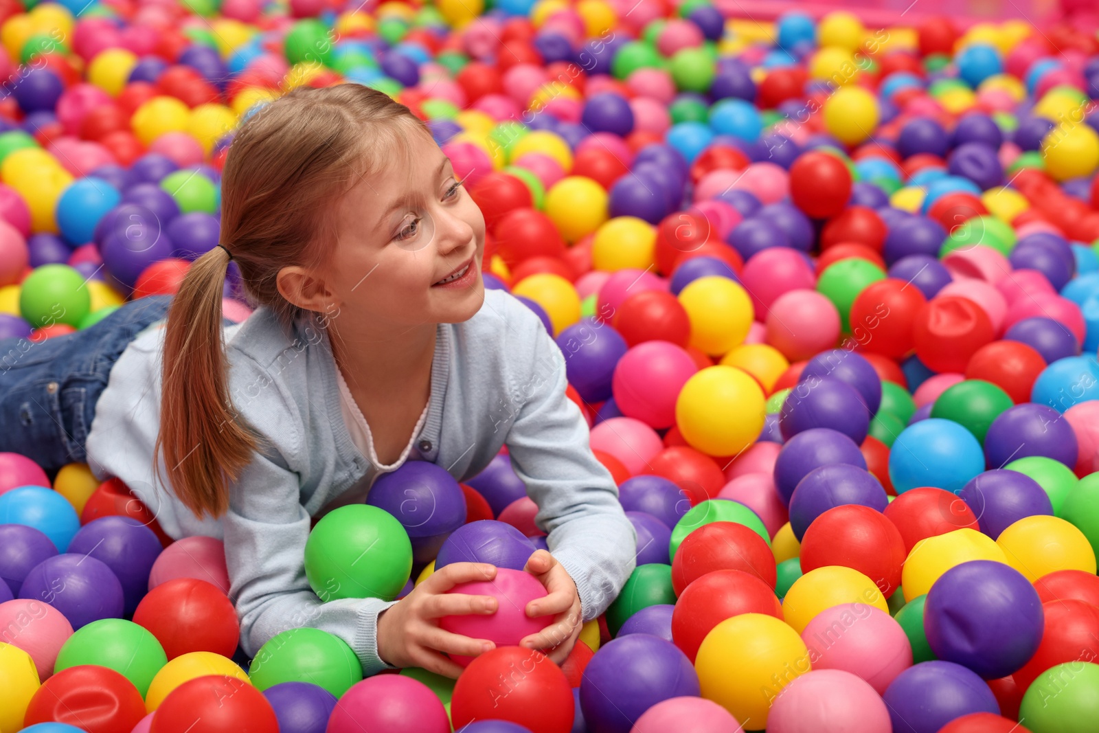 Photo of Happy little girl lying on many colorful balls in ball pit, space for text