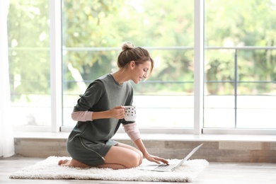 Photo of Young woman with cup of aromatic coffee and laptop sitting on floor near window at home