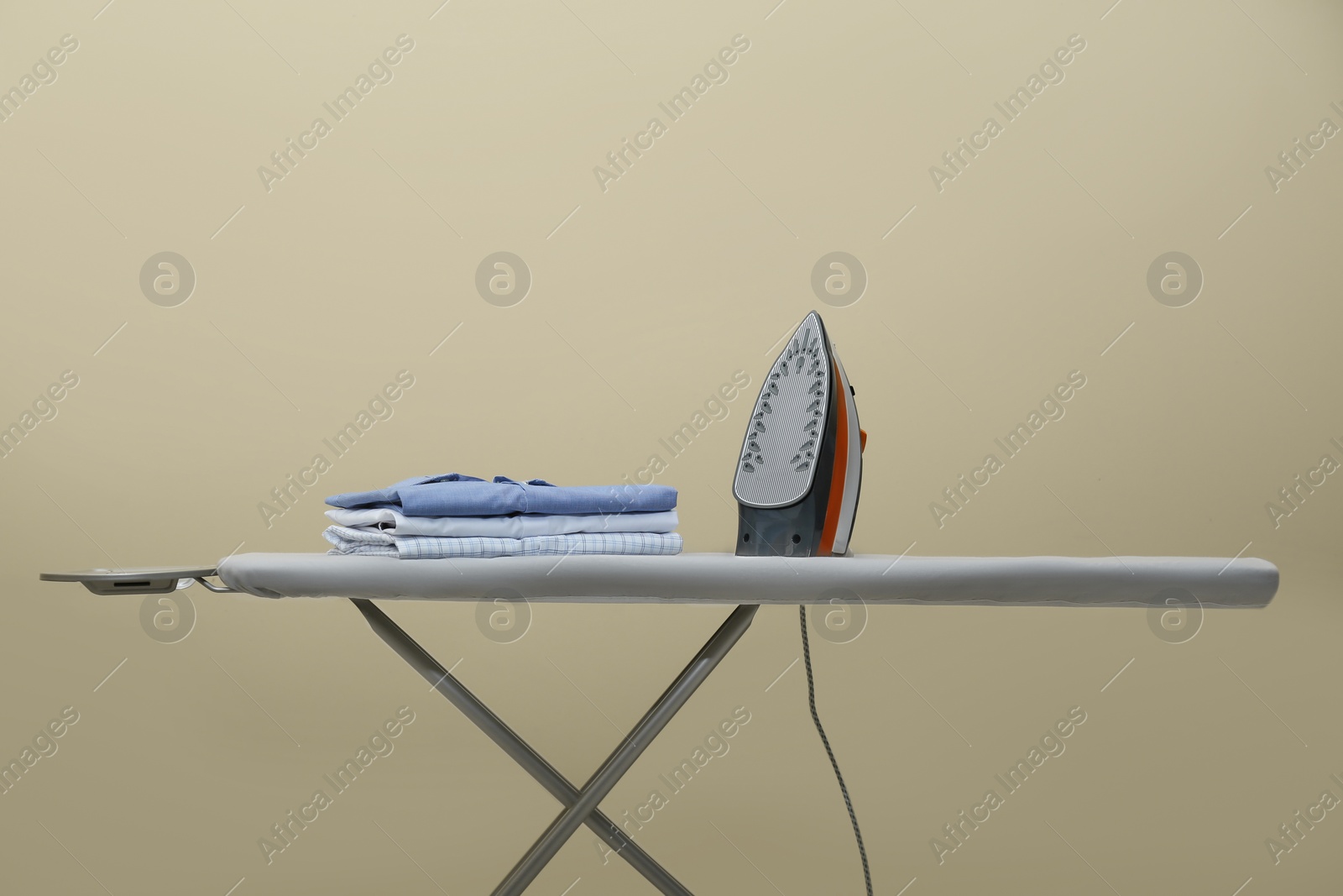 Photo of Modern iron and stack of clean clothes on board against beige background