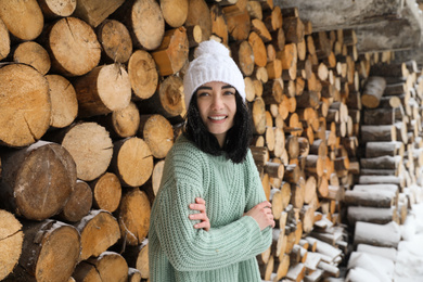 Young woman wearing warm sweater and hat near stack of firewood outdoors. Winter season
