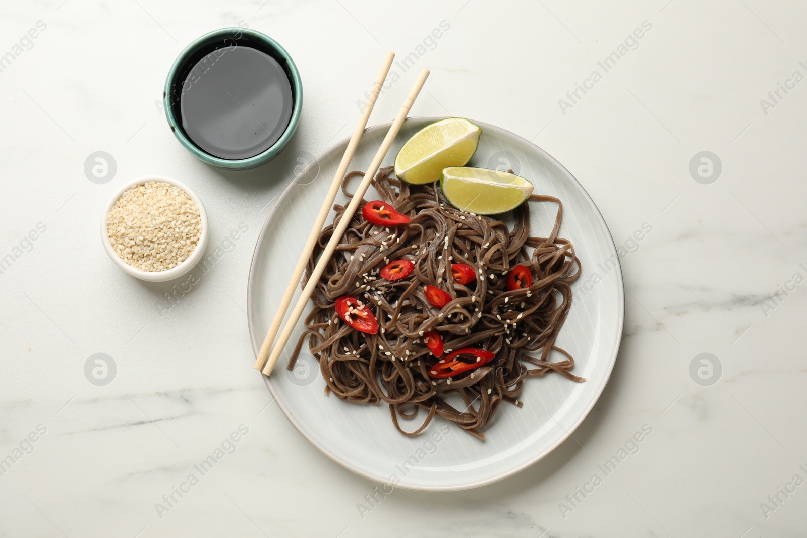 Photo of Tasty buckwheat noodles (soba) with chili pepper and lime served on white marble table, flat lay