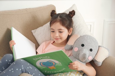 Photo of Little girl with toy elephant reading book in armchair at home