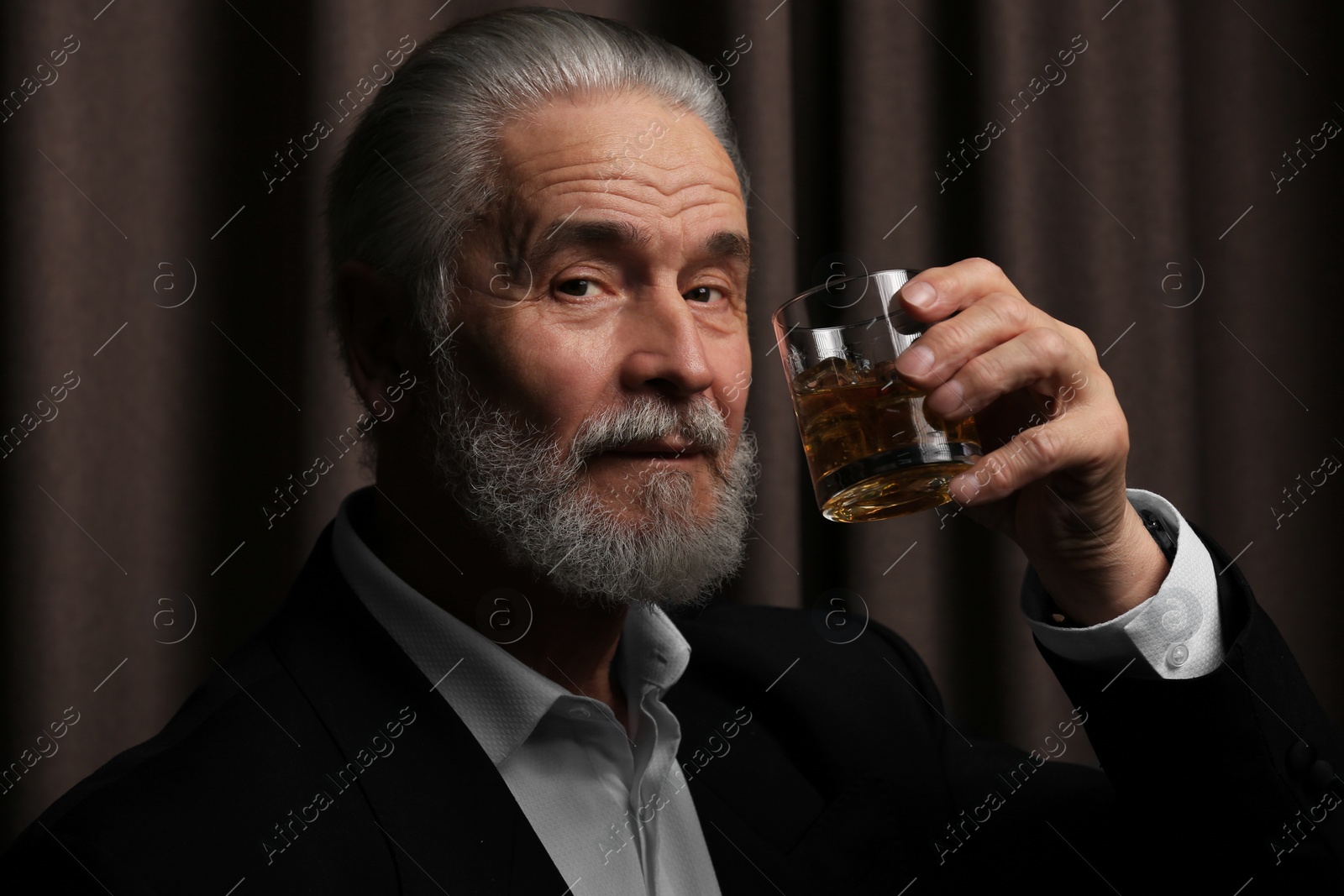 Photo of Senior man in formal suit holding glass of whiskey with ice cubes on brown background