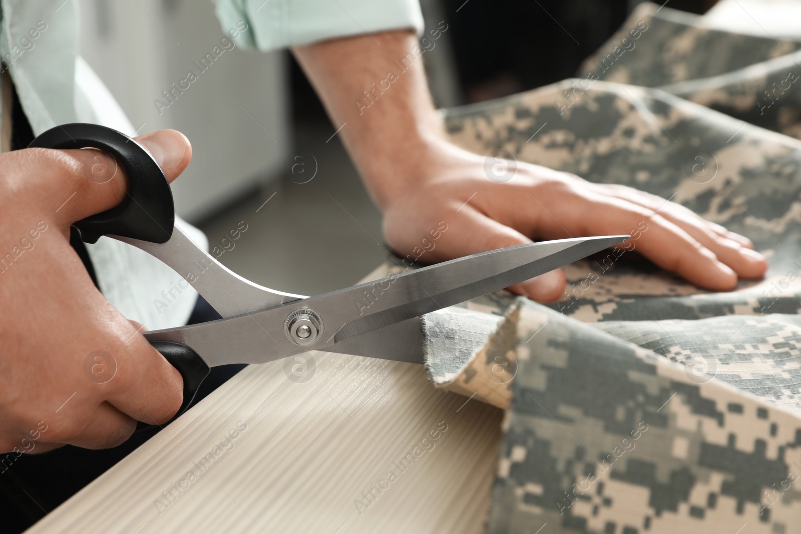 Photo of Professional tailor cutting camouflage fabric with scissors in workshop, closeup