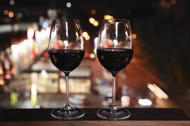 Photo of Glasses of red wine on wooden surface against blurred cityscape. Modern outdoor terrace