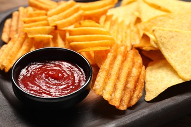 Photo of Tasty ridged and tortilla chips with ketchup on wooden tray, closeup