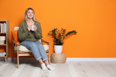 Young woman with cup of drink relaxing in armchair at home, space for text. Interior design