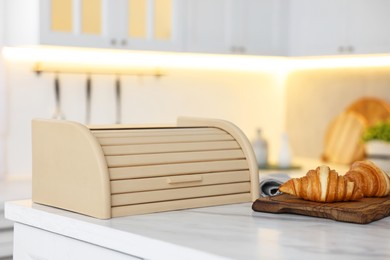 Photo of Wooden bread box and board with croissants on white marble table in kitchen