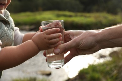 Photo of Father giving her daughter glass of fresh water near stream on sunny day, closeup