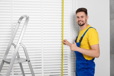 Photo of Worker in uniform using measuring tape while installing horizontal window blinds near stepladder indoors