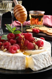 Photo of Pouring honey onto brie cheese served with raspberries and walnuts on black plate, closeup