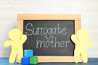 Small chalkboard with phrase Surrogate Mother, paper people cutouts and cubes on blue wooden table