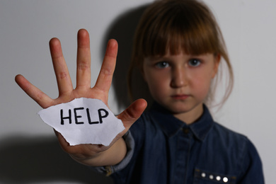 Abused little girl with sign HELP near white wall, focus on hand. Domestic violence concept