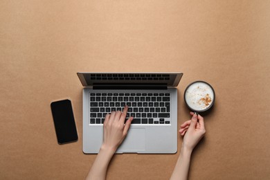 Woman with cup of cappuccino using laptop at light brown table, top view. Space for text