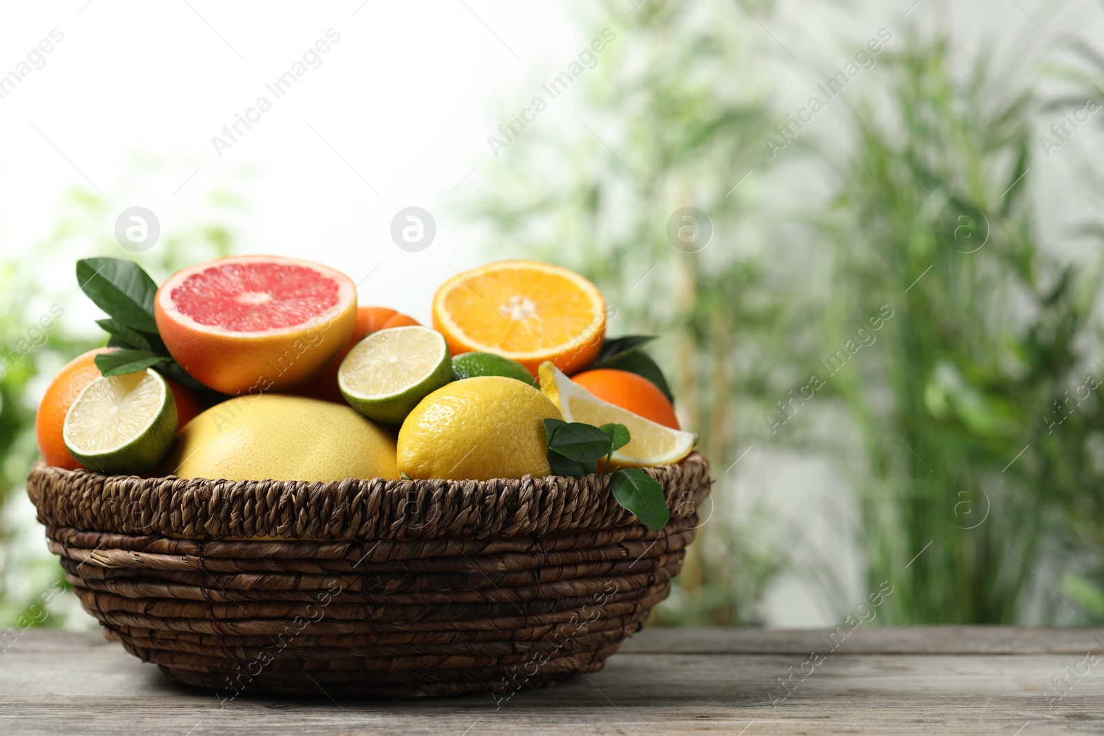 Photo of Different fresh citrus fruits and leaves in wicker basket on wooden table against blurred background, closeup. Space for text