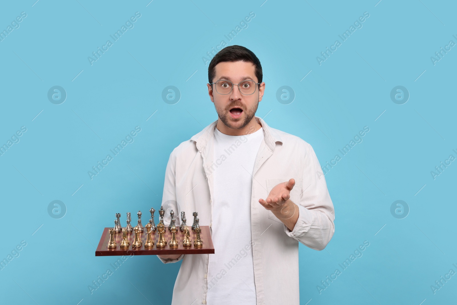 Photo of Emotional man holding chessboard with game pieces on light blue background