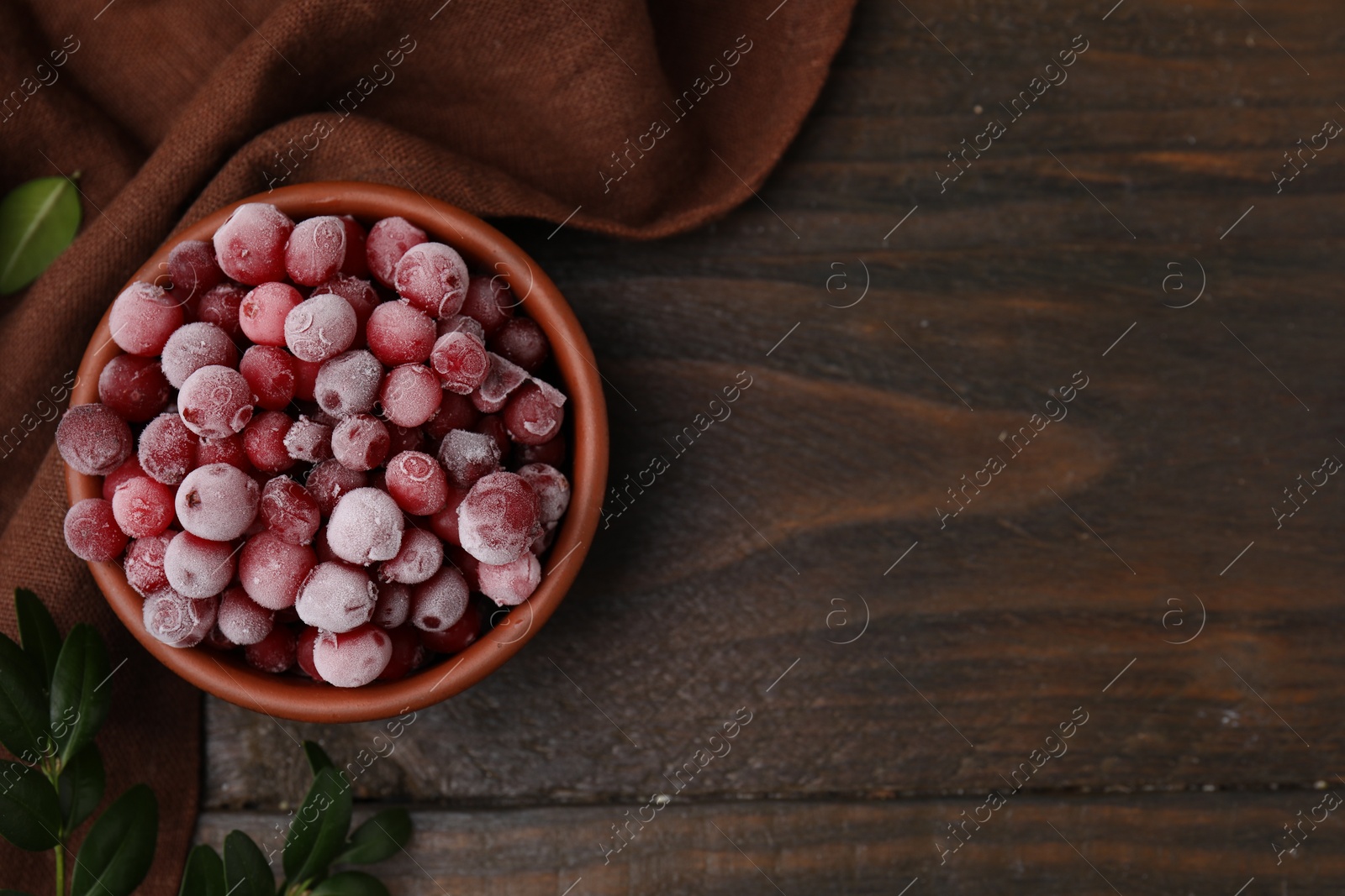 Photo of Frozen red cranberries in bowl and green leaves on wooden table, top view. Space for text