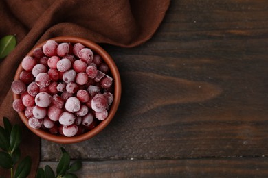 Photo of Frozen red cranberries in bowl and green leaves on wooden table, top view. Space for text