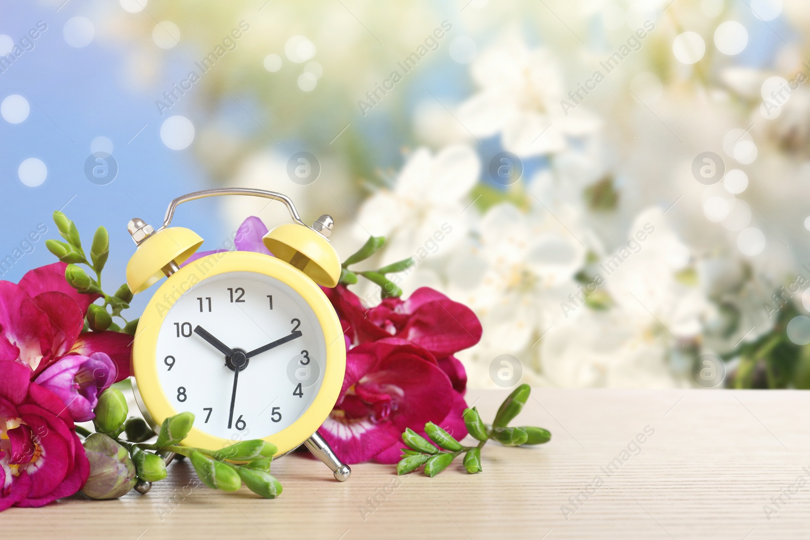 Image of Alarm clock and flowers on wooden table against blurred background, space for text. Spring time