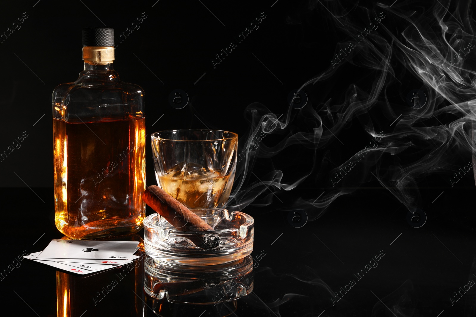 Photo of Smoldering cigar, ashtray, playing cards and whiskey on black mirror surface. Space for text