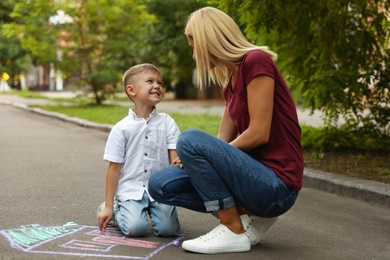 Photo of Nanny with cute little boy drawing house with chalks on asphalt