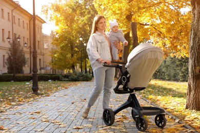 Photo of Happy mother walking with her baby daughter and stroller outdoors on sunny autumn day, space for text