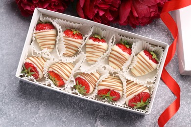 Photo of Delicious chocolate covered strawberries in box and flowers on light grey table, flat lay