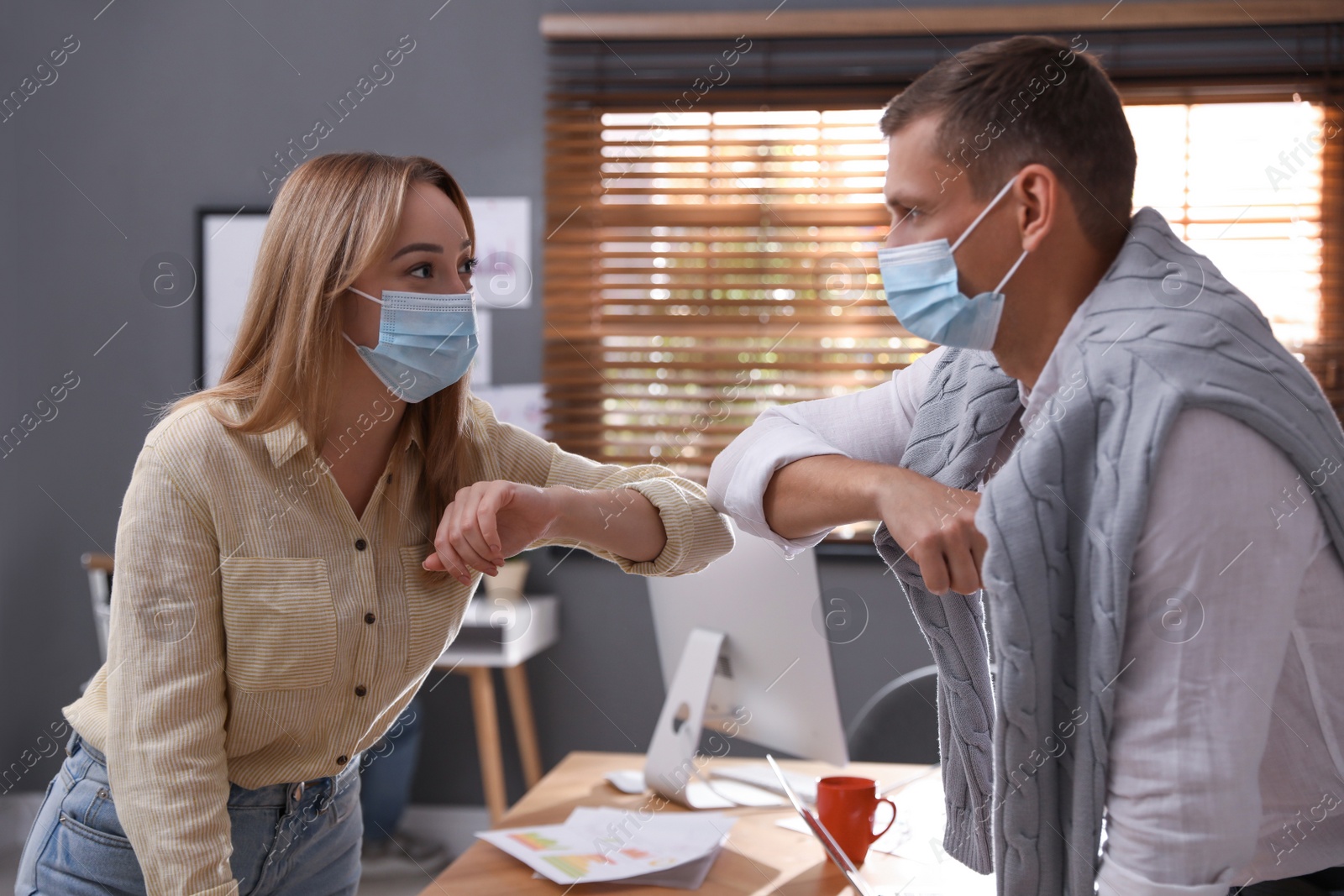 Photo of Coworkers with protective masks making elbow bump in office. Informal greeting during COVID-19 pandemic