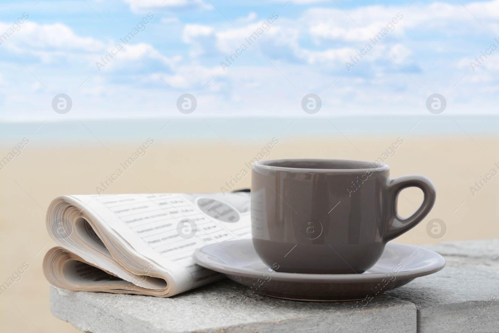 Photo of Ceramic cup of hot drink and newspaper on stone surface near sea in morning