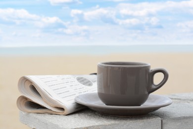 Ceramic cup of hot drink and newspaper on stone surface near sea in morning