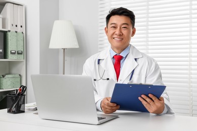 Photo of Happy doctor with laptop and clipboard consulting patient at white desk in clinic. Online medicine