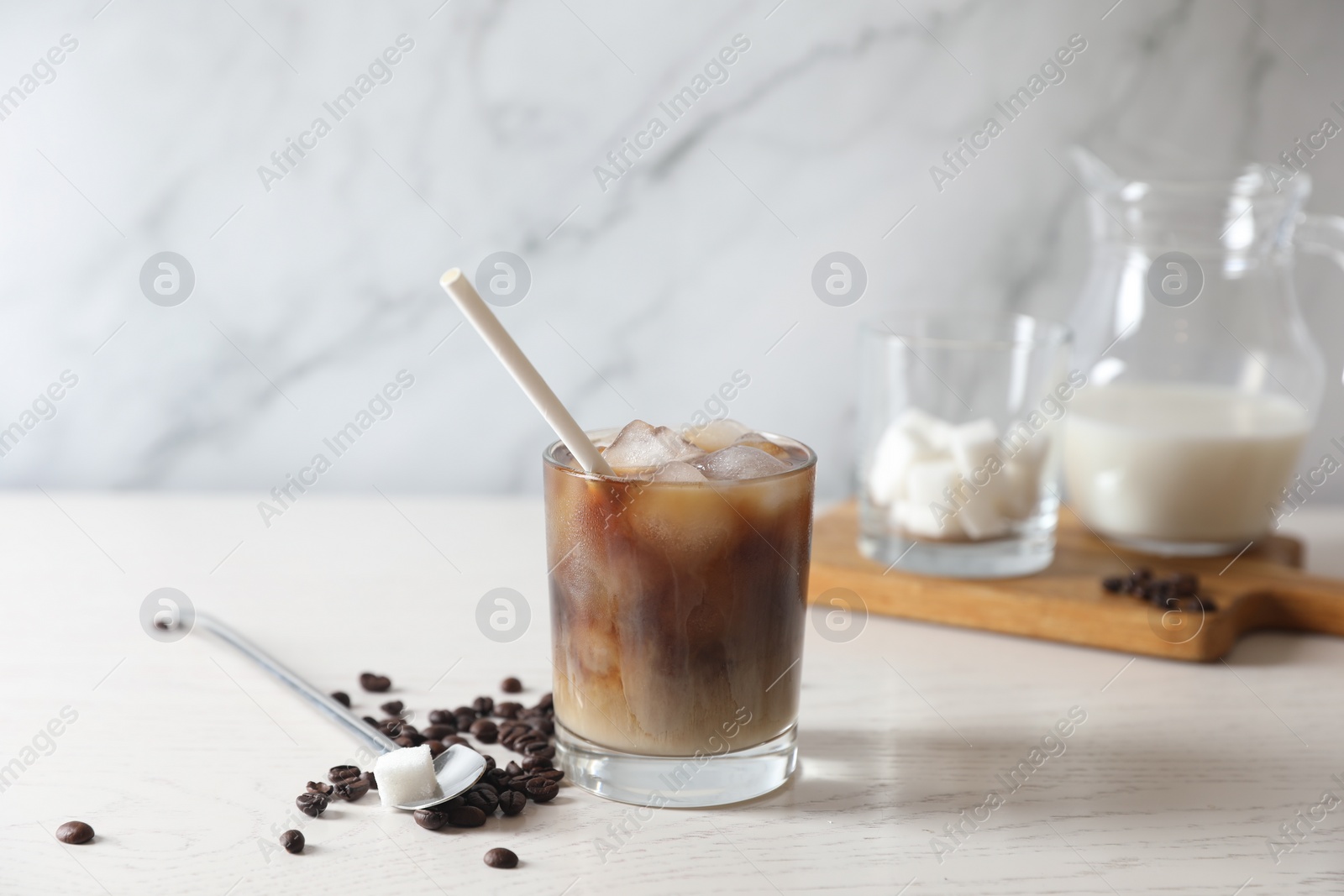 Photo of Refreshing iced coffee with milk in glass, beans, spoon and sugar cube on white wooden table