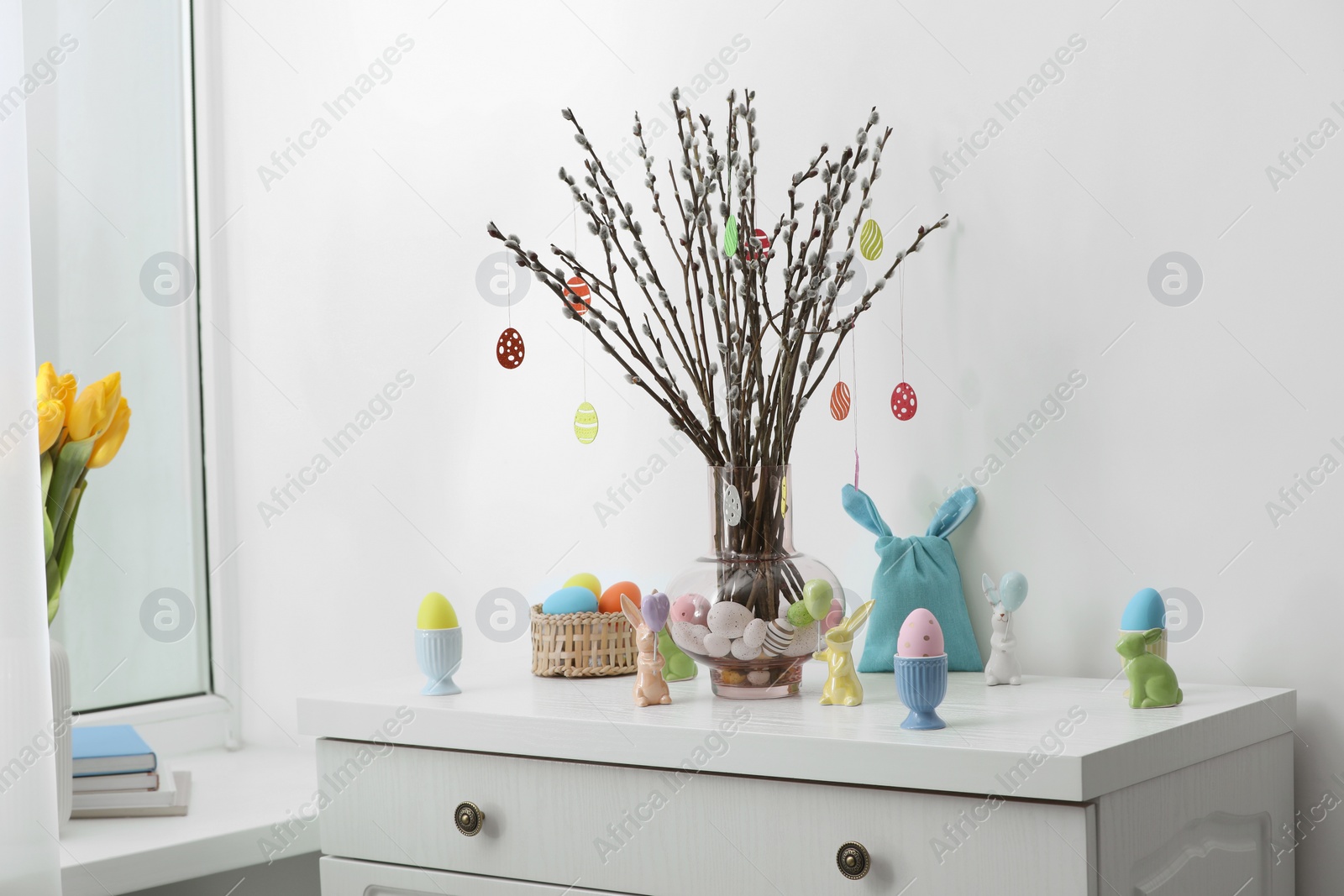 Photo of Beautiful pussy willow branches with paper eggs in vase and Easter decor on white chest of drawers at home