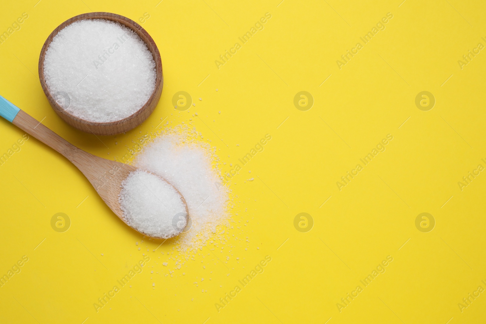 Photo of Bowl and spoon with granulated white sugar on yellow background, flat lay. Space for text