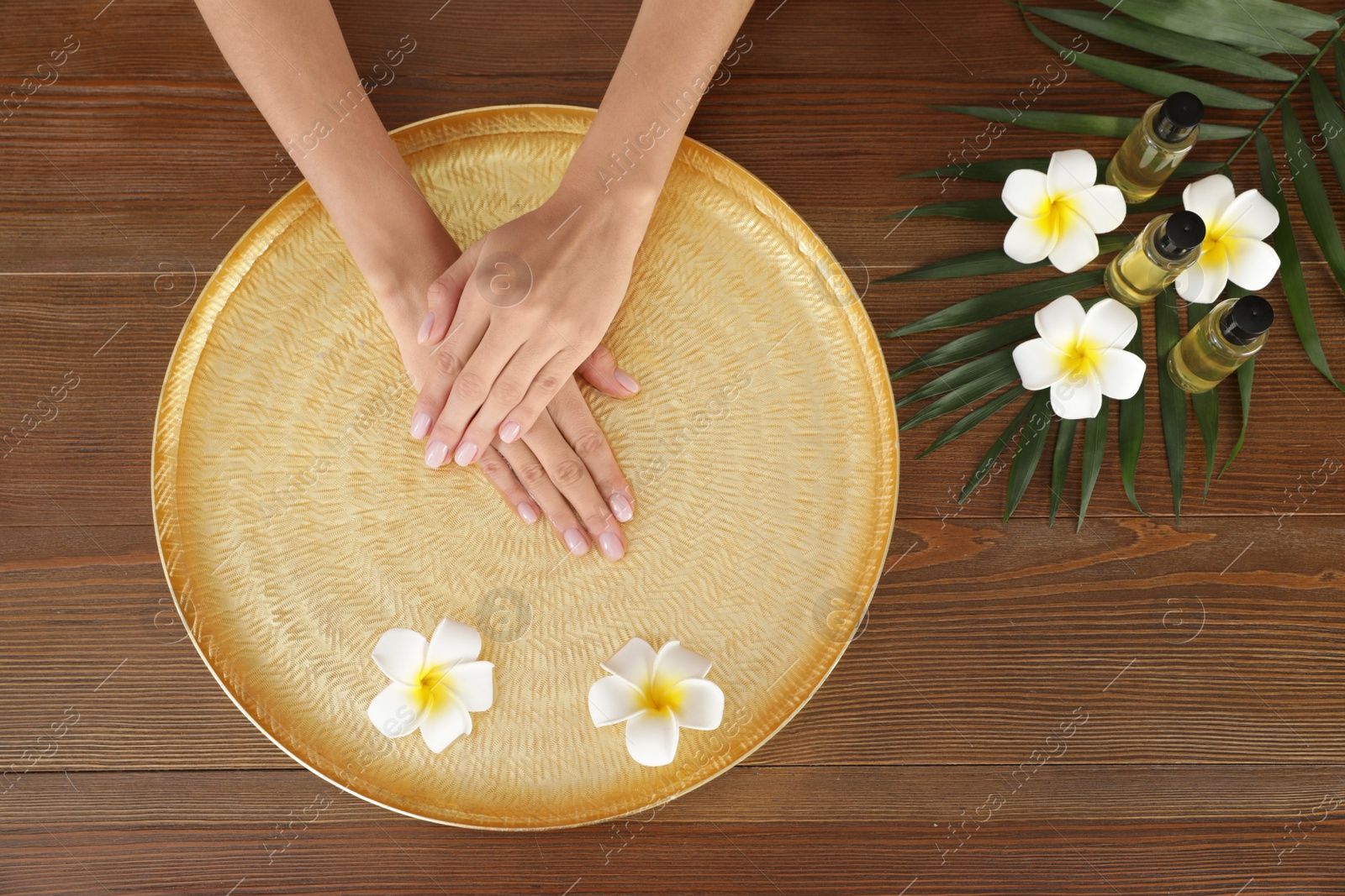 Photo of Woman soaking her hands in bowl with water and flowers on wooden table, top view. Spa treatment