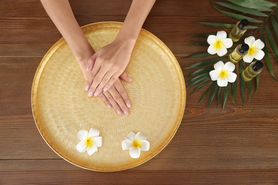 Photo of Woman soaking her hands in bowl with water and flowers on wooden table, top view. Spa treatment