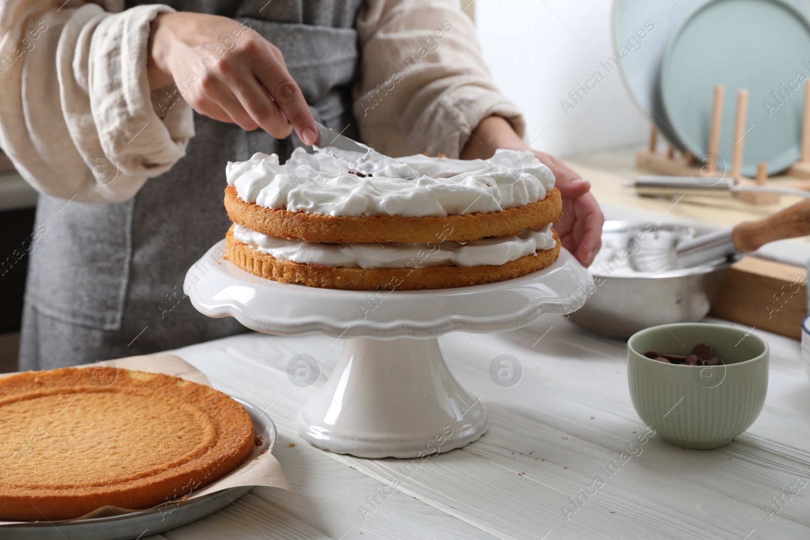 Photo of Woman smearing sponge cake with cream at white wooden table in kitchen, closeup