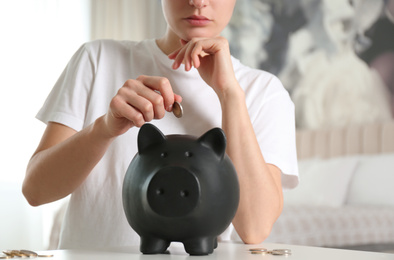 Photo of Woman putting money into piggy bank at white table indoors, closeup