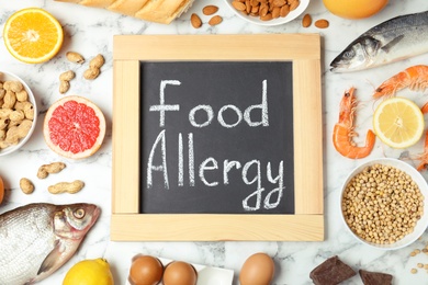 Chalkboard with words FOOD ALLERGY and different products on white marble table, flat lay