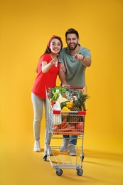 Photo of Young couple with shopping cart full of groceries on yellow background